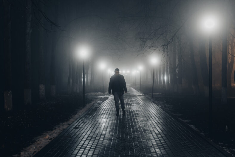 Sad man alone walking along the alley in night foggy park. front view.