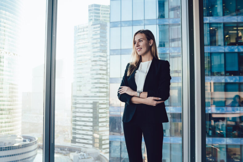 Smiling blonde woman in trendy suit posing in office interior near window with panoramic view on business center, cheerful prosperous female proud ceo in formal outfit standing with crossed hands