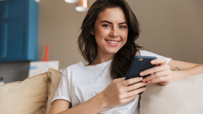 Beautiful smiling young brunette woman relaxing on a couch at home, using mobile phone
