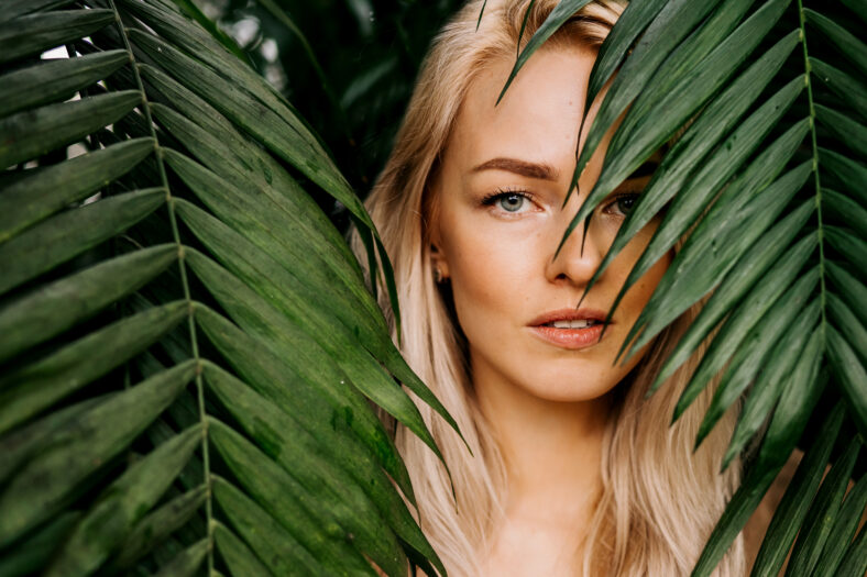 Young woman's face surrounded by tropical leaves. Closeup portrait of magnificent lady with natural nude makeup, perfect skin, blonde hair. Natural cosmetic, wellness, purity, skincare, beauty concept
