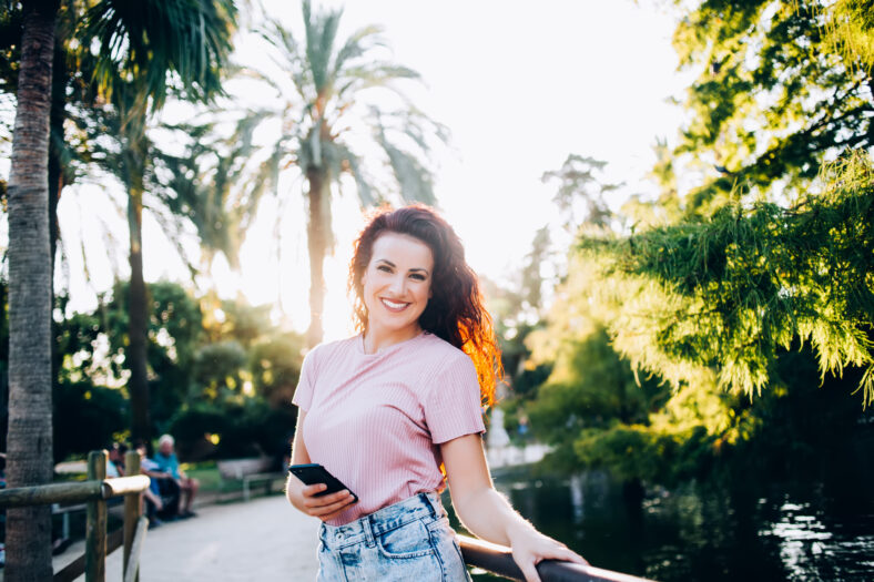 Portrait of cheerful curly brunette woman enjoying sunny day recreation in park on summer vacation holding mobile phone, smiling female blogger looking at camera using smartphone for share content