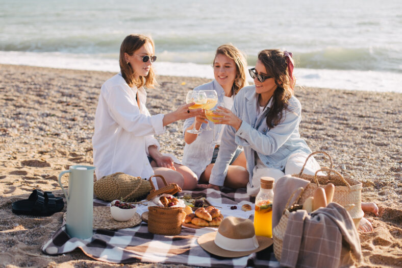 Three young beautiful happy women female friends having cozy summer picnic with lemonade, fresh bread and fruits on a beach at sunset.
