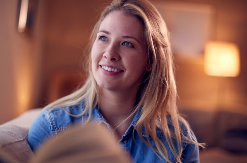 Woman At Home Relaxing And Reading Book Sitting On Sofa In Evening