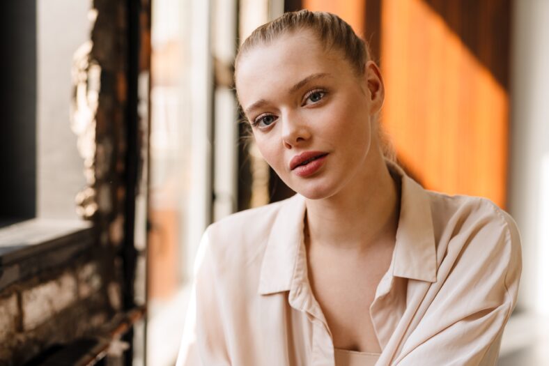 Young blonde woman dressed in shirt posing and looking at camera indoors