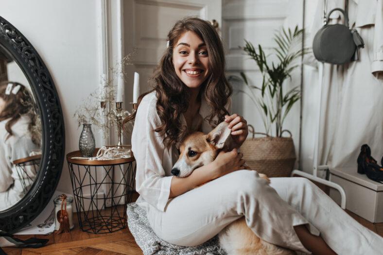 Curly woman in white shirt having fun with dog. Brunette pretty lady in beige blouse and pants plays and hugs corgi while sitting on floor