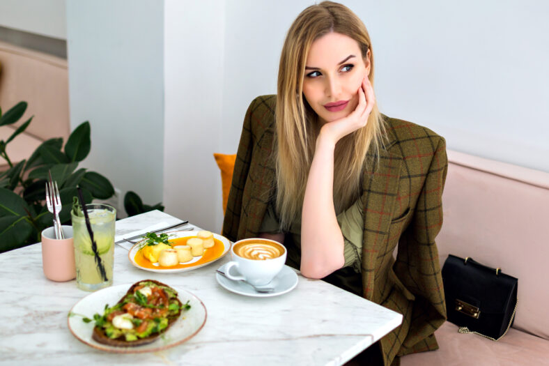 Lifestyle indoor portrait of stylish young blonde business woman enjoying her brunch at hipster cafe, asking for waiter and smiling, co working space.
