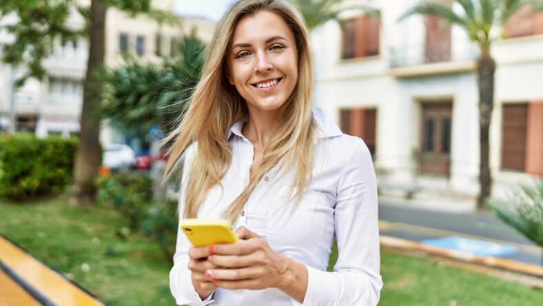 Beautiful blonde woman smiling happy outdoors on a sunny day using smartphone