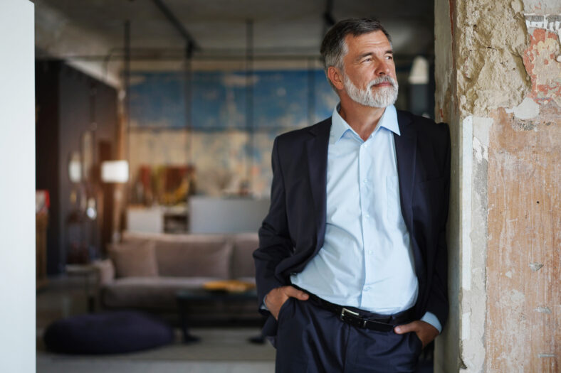 Positive confident mature executive man wearing black suit and blue shirt smiling and looking away while standing in office