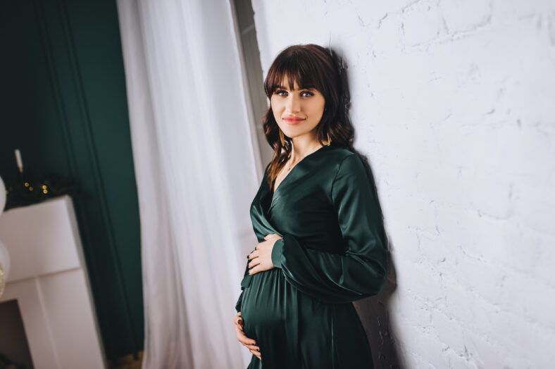 A beautiful pregnant brunette girl in a green dress with a big belly stands near a white wall in the interior.