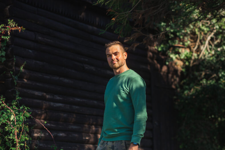 Handsome bearded man in a green sweater posing by log wall and green trees on the sunset. Young eco friendly hipster.
