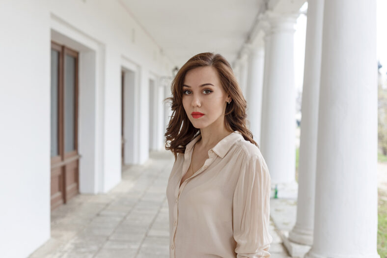 pretty caucasian young woman in beige blouse, red lipstick and wavy hair standing on veranda of cafe. white concrete pillars in background