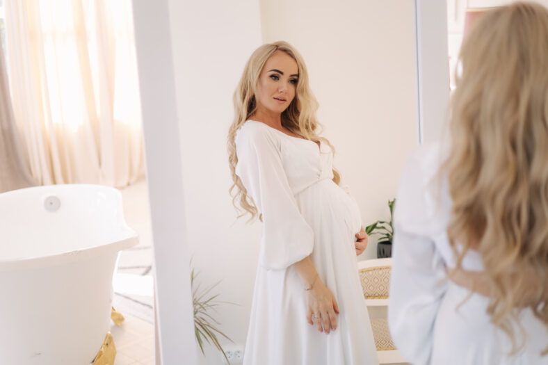 Beautiful portrait of pregnant woman in white dress. Concept of perfect pregnancy. Woman put her hand on belly. Future mother is waiting for baby.