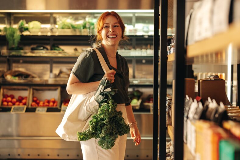 Female customer smiling cheerfully while carrying a bag with fresh organic vegetables in a grocery store. Happy young woman grocery shopping in a trendy supermarket.