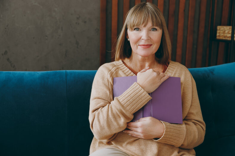 Smiling happy elderly woman 50s years old wearing casual clothes glasses sit on blue sofa read book novel look camera stay at home flat rest relax spend free spare time in living room indoor grey wall