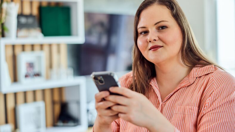 A pretty overweight woman sitting in the living room at home, holding her mobile phone and looking at the camera.
