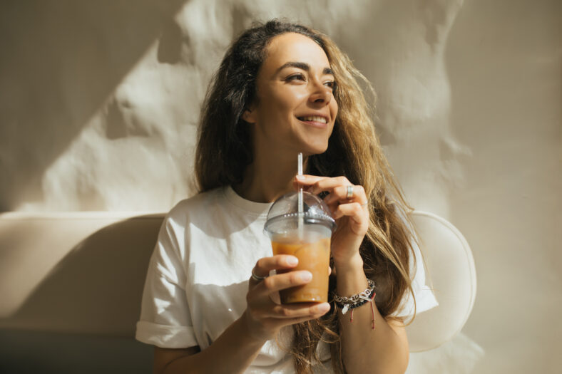 Young beautiful woman with long curly hair drinking iced coffee on beige background.