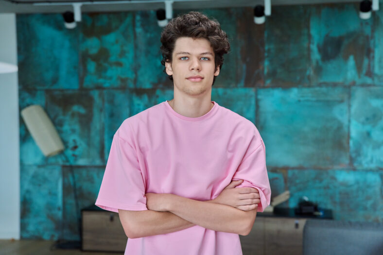 Portrait of a handsome guy 18, 19 years old with crossed arms in home interior. Curly confident young college student in pink t-shirt looking at camera. Youth, age, lifestyle concept