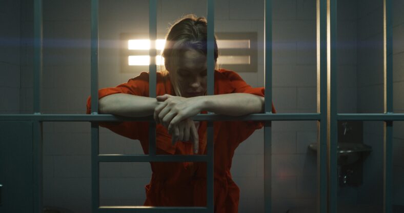 Female prisoner in orange uniform holds hands on metal bars, walks, looks at barred window in jail cell. Woman serves imprisonment term for crime in prison. Depressed inmate in correctional facility.