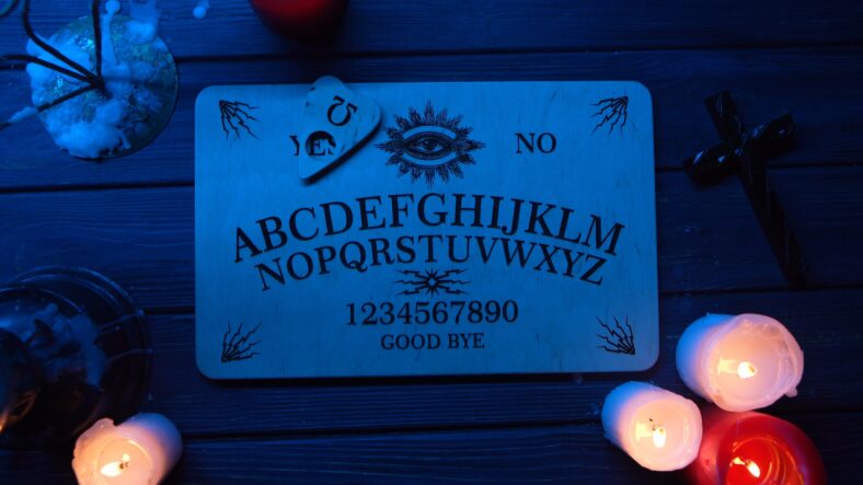 Close up shot of a wooden ouija board with a planchette placed on a table. There are candles, book, cross and cantlesticks around. The light is muffed, blue. Creative mystical content.