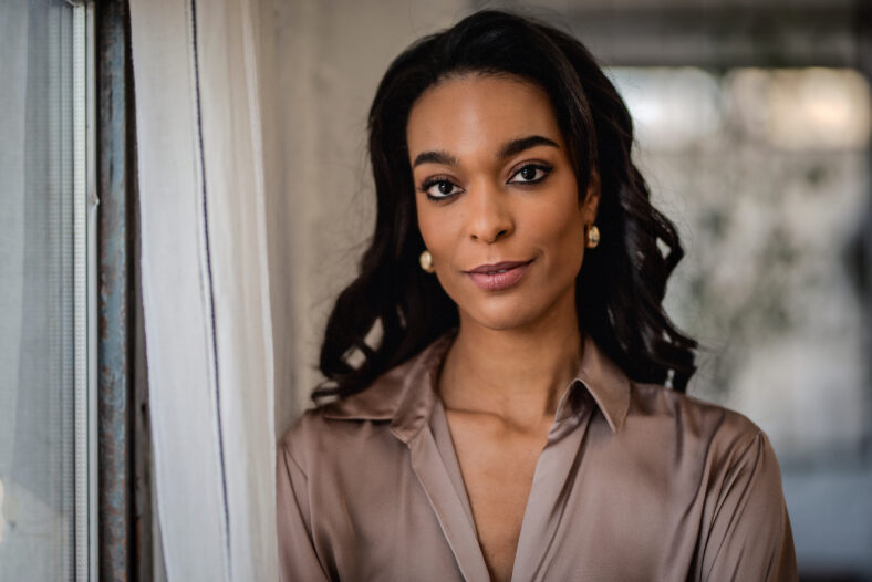 Close-up portrait of a young african-american woman standing at the window and looking at camera. Long haired female wearing casual clothes.