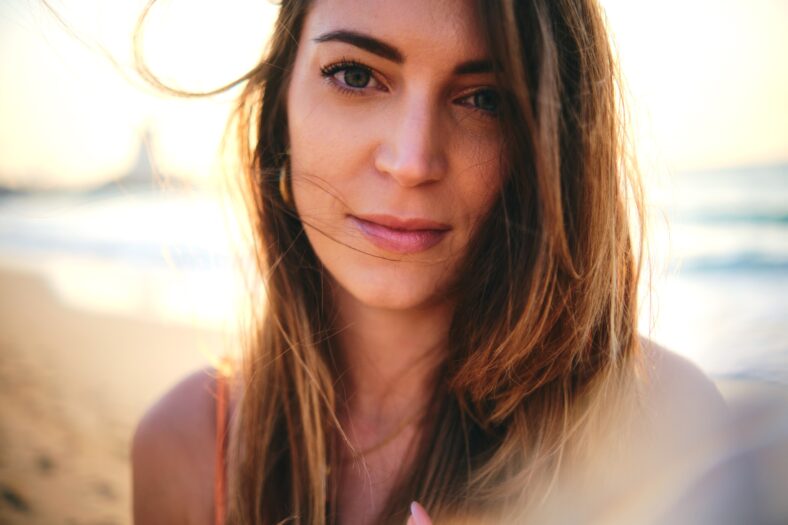 An intimate close-up image of a woman's face with her hair blowing in the wind, on the beach during sunset