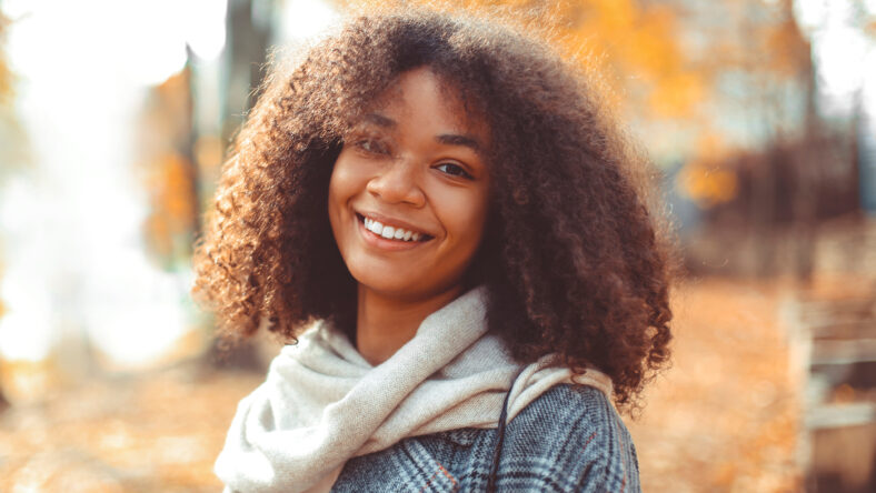 Cute autumn close up portrait of young smiling happy african american woman with curly hair enjoying walk in park in fall season. Outdoor photo of pleased black girl with eyes in love with life