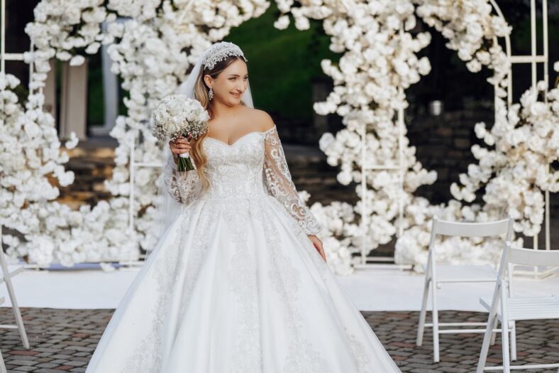 A woman in a white wedding dress is standing in front of a white archway. She is holding a bouquet of flowers and she is posing for a picture. Concept of elegance and celebration