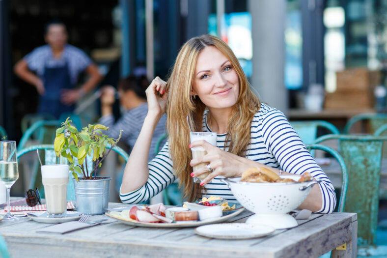 Happy beautiful woman drinking coffee with milk and having healthy breakfast in outdoor cafe in summer city in Europe.