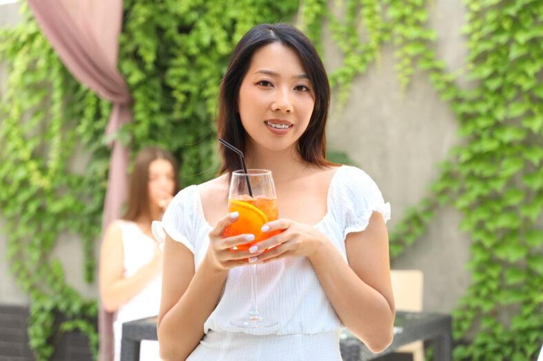 Beautiful young Asian woman with glass of tasty aperol spritz cocktail in cafe, outdoors