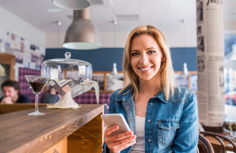 Blond woman in denim shirt sitting at the bar in modern city cafe, holding a smart phone, texting