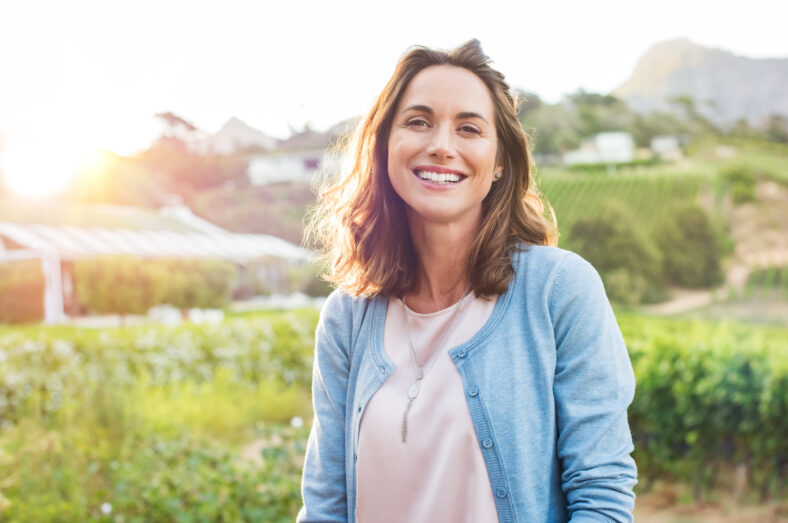 Cheerful mature woman enjoying in park during sunset. Happy hispanic woman in casual feeling relaxed outdoor. Smiling brunette woman looking at camera with carefree.