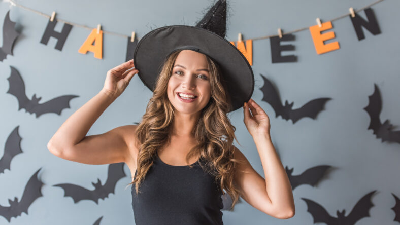 Beautiful young woman in witch hat is looking at camera and smiling, on background decorated for Halloween