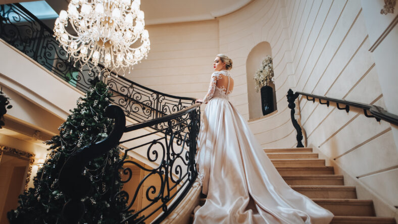 Beautiful bride in a white dress with long train is climbs up the stairs in a classic interior.