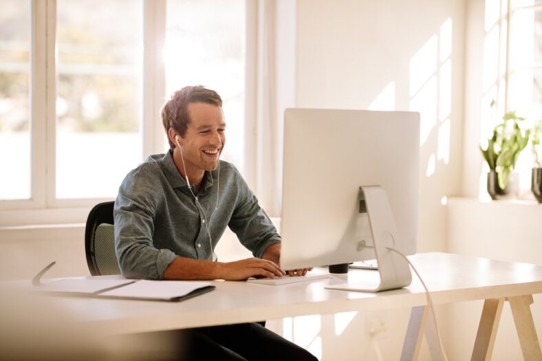 Smiling businessman working on computer. Man sitting at his work table working on computer at home listening to audio using earphones.