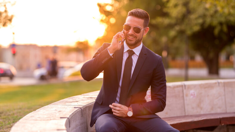 A handsome young businessman sitting on a bench and talking on his phone on a sunny day.