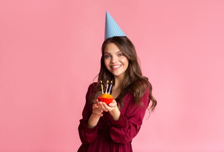 Happy young woman in party hat holding birthday cupcake with lighted candles over pink studio background, empty space