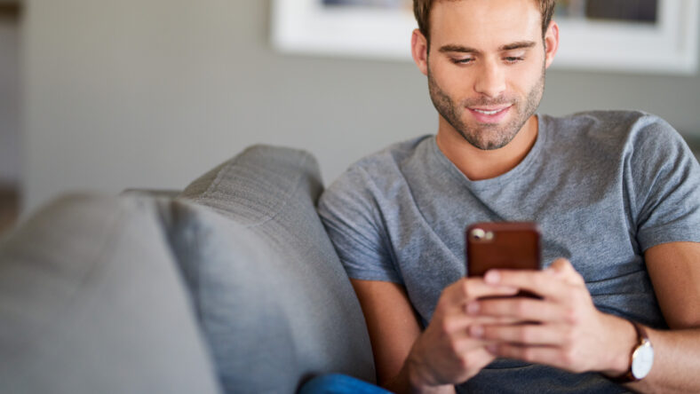 Young man texting on his cellphone and smiling while relaxing alone on his living room sofa at home