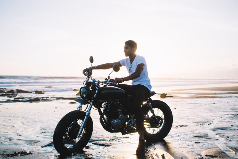 Side view of Hispanic adult man in white shirt and black pants looking ahead while sitting on motorcycle and holding handlebar on sand ocean coast during sunrise on blurred background