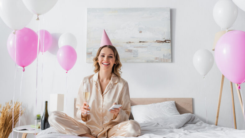 Smiling woman with smartphone and champagne celebrating birthday in bedroom