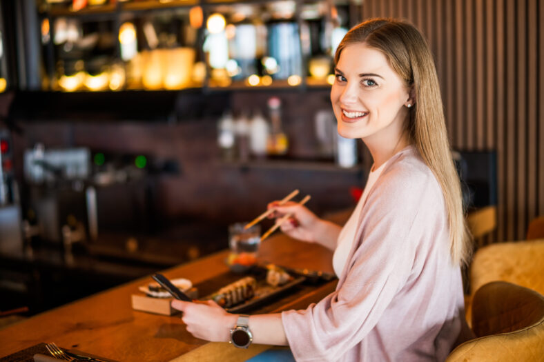 Happy woman is eating sushi with chopsticks at sushi restaurant.
