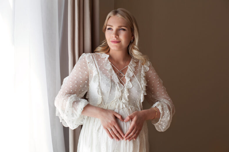 Charming blonde pregnant woman stands near window and makes heart sign with her fingers, looks away, wears white light cotton dress