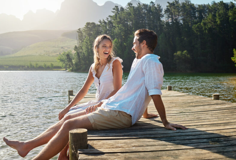 Love, laugh and pier with couple at lake for bonding, romance and affectionate date. Nature, travel and holiday with man and woman sitting on boardwalk in countryside for happy, summer and vacation.