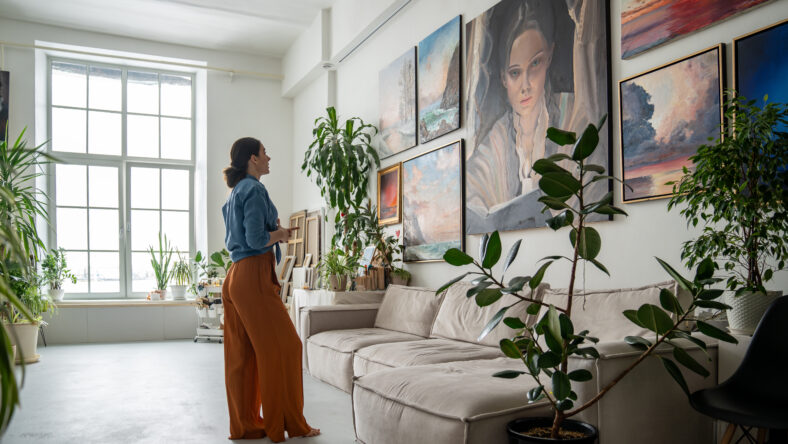 Female artist deeply reflects on portrait amidst other artworks on wall in studio. Creative workshop space with diverse collection of paintings, woman painter inspiration and new ideas for work
