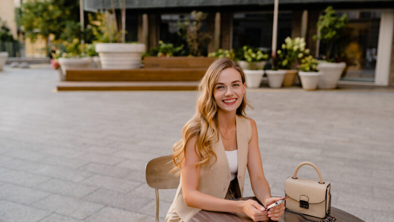 attractive young blond woman sitting in cafe using smart phone drinking coffee in spring summer autumn fashion trend outfit smiling happy wearing beige suit street style, chic glam make-up accessories and hair