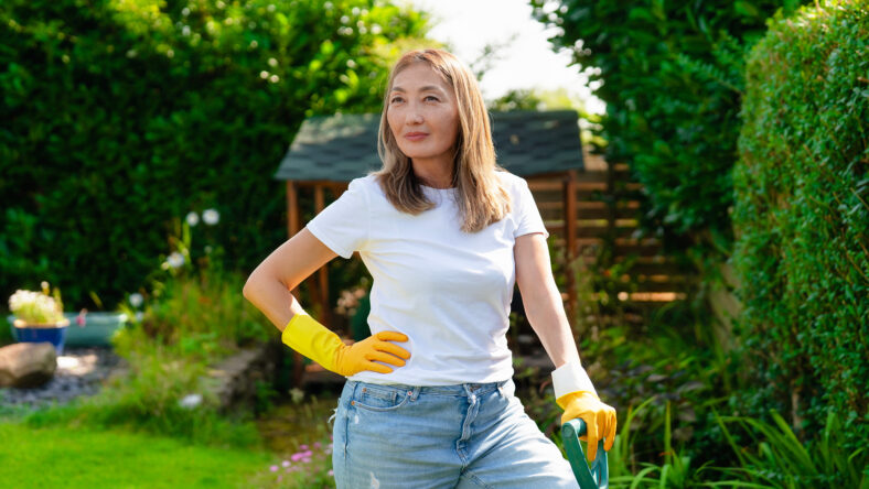 Woman gardening in sunny backyard wearing gloves and casual clothes, enjoying a day of outdoor work and nature in a lush green environment