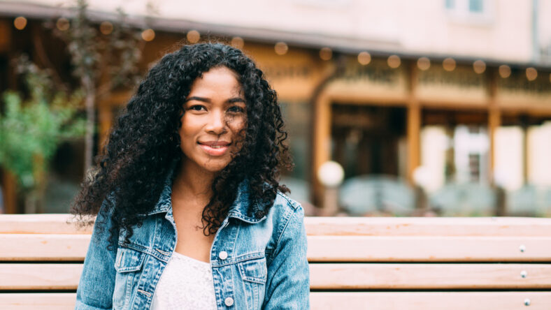 Portrait of pretty young african american woman in blue jeans jacket sitting on wooden bench outdoor.
