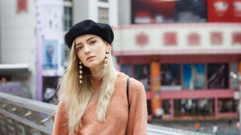 A beautiful young blonde woman dressed in a large warm pink sweater with a black beret and gorgeous earrings. She goes through a big spectacular Chinese city in autumn.