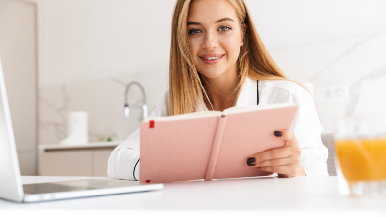 Photo of smiling pretty woman in pajama making note in diary and drinking juice while sitting at table in bright kitchen