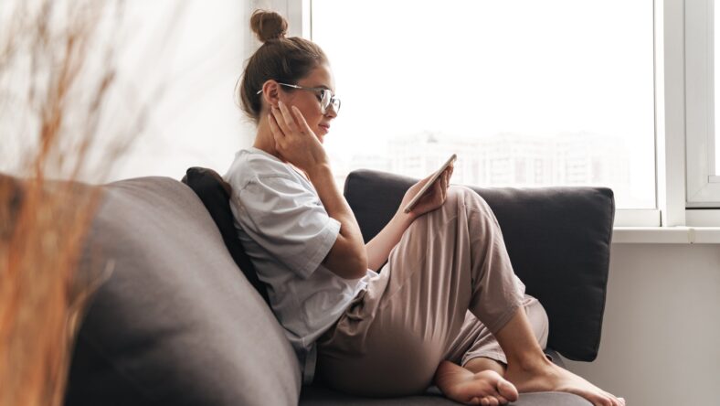 Image of concentrated nice woman in eyeglasses using cellphone while sitting on sofa at living room