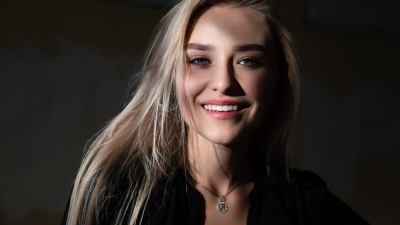 Happy smiling blonde girl with healty natural skin, dressed in black dress, stands in the shade, the rays of the sun fall on her face, outdoor photo